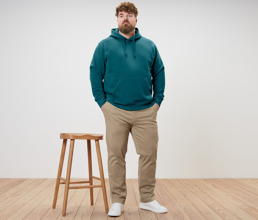 Mężczyzna stoi z rękami w kieszeniach. Ma na sobie ciemnoturkusową bluzę z kapturem i beżowe spodnie chinosy.