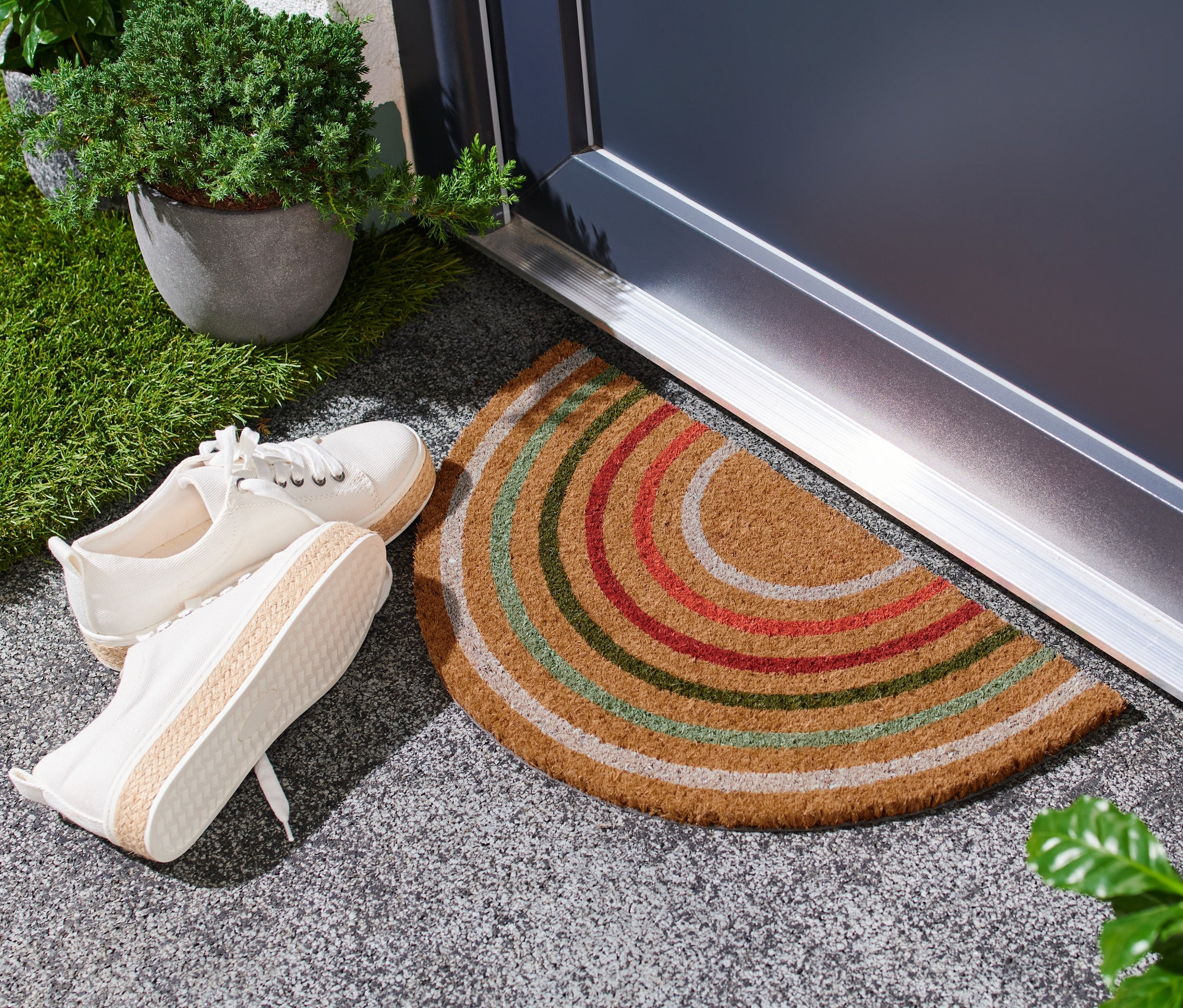 Przed drzwiami leży wycieraczka kokosowa z kolorowymi paskami, obok stoi para białych butów.