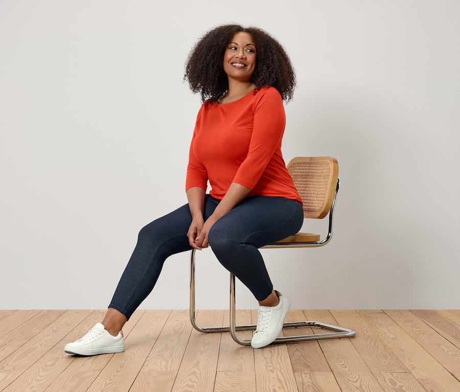 Kobieta siedzi na krześle, ubrana w pomarańczową bluzkę, granatowe legginsy i białe trampki.