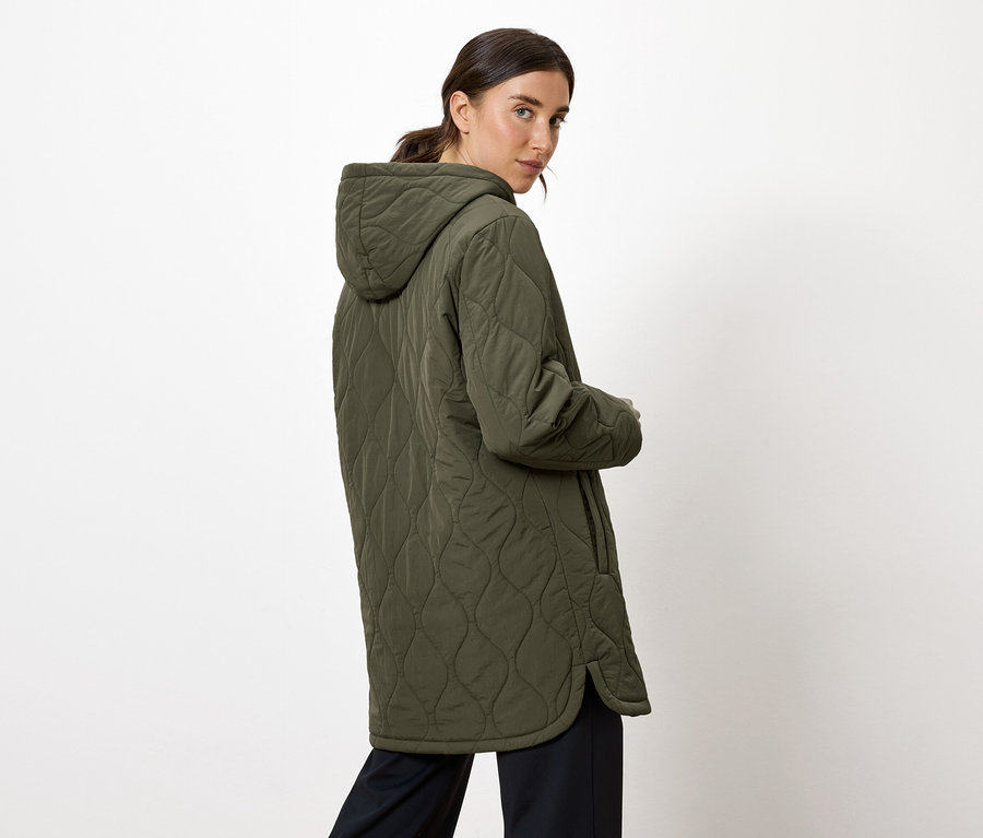 Kobieta spogląda przez ramię, ubrana w oliwkową kurtkę z kapturem.