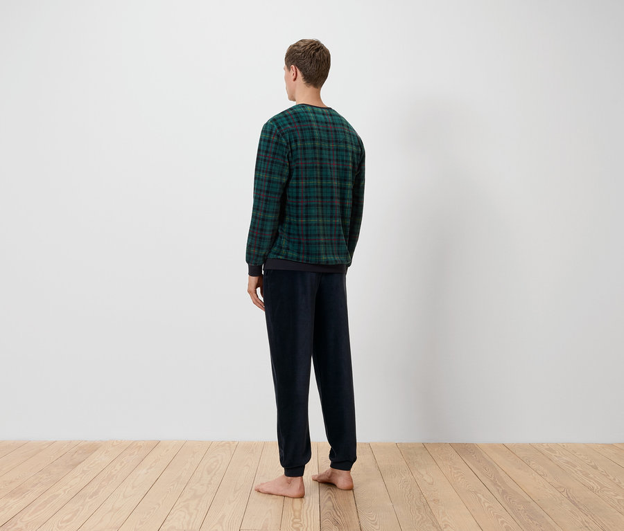 Mężczyzna tyłem do kamery ma na sobie zielono-czarną koszulę w kratę i granatowe spodnie, stoi na drewnianej podłodze przed białą ścianą.
