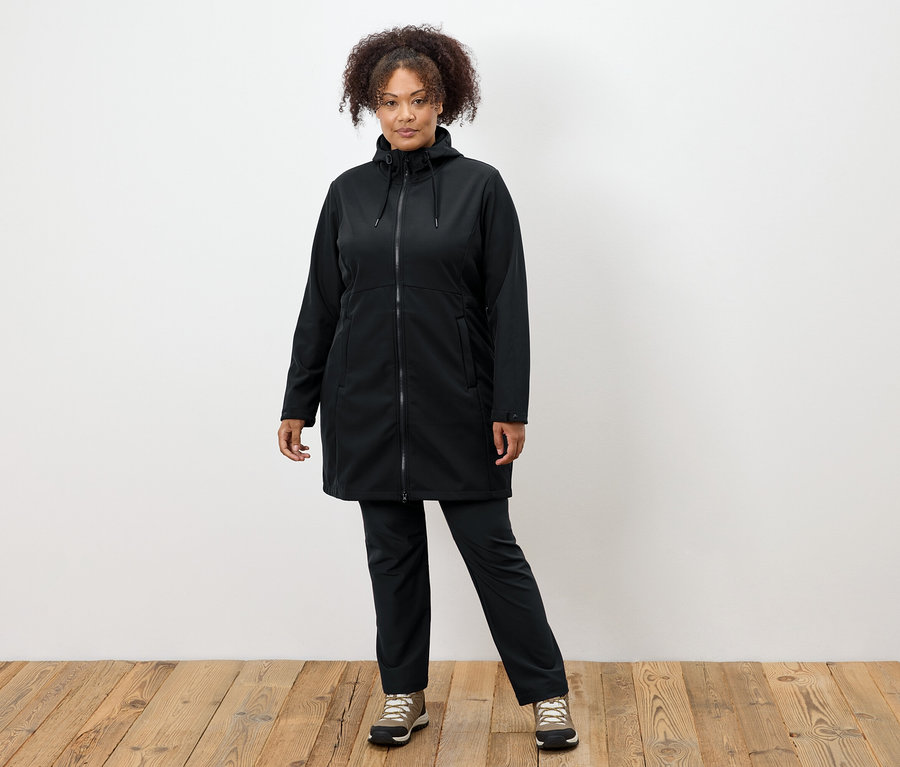 Kobieta w czarnym płaszczu i spodniach stoi na drewnianej podłodze.