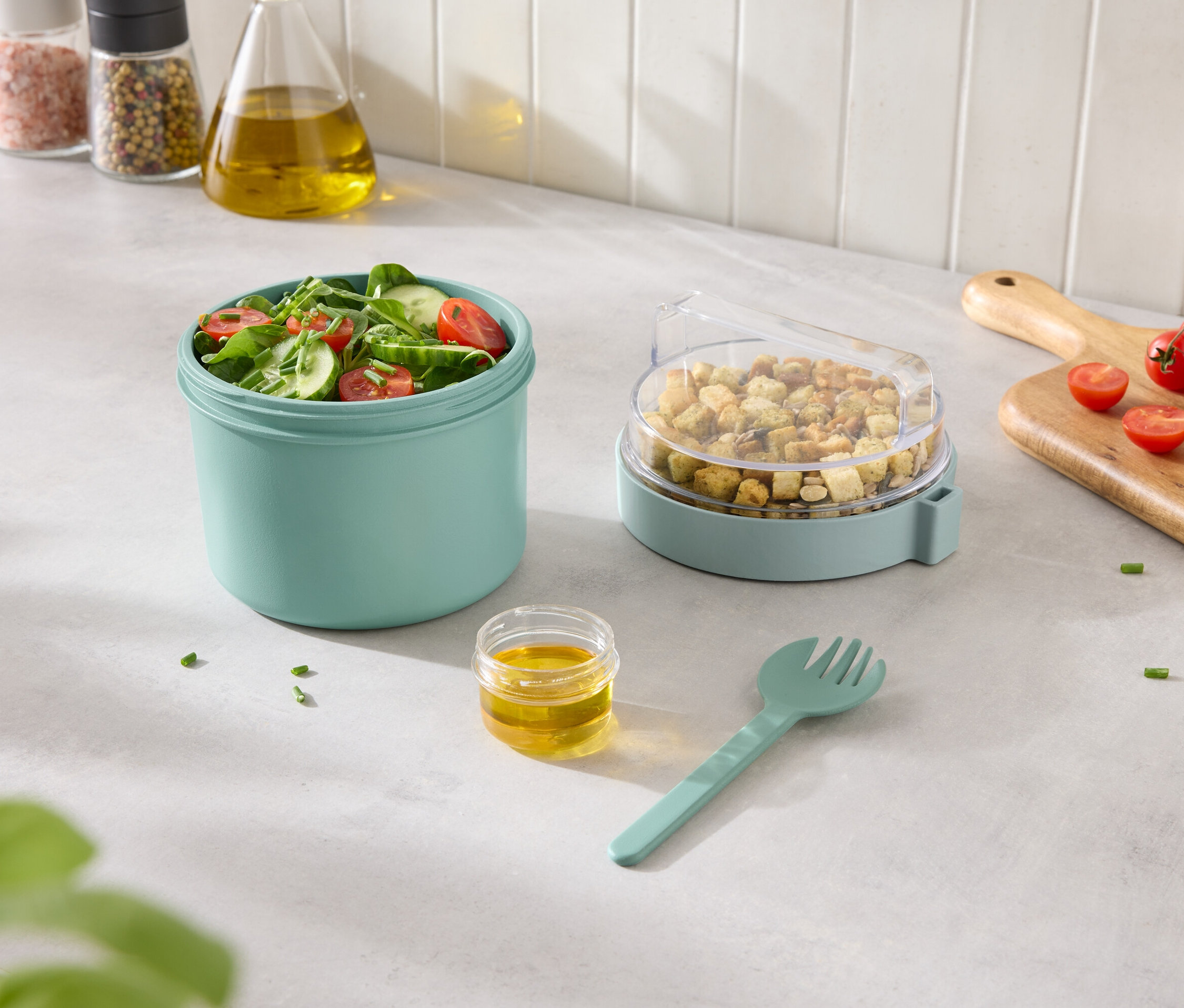 Sałatka warzywna w turkusowym pojemniku z grzankami w pojemniku z przezroczystą pokrywką oraz dressingiem i widelcem obok.