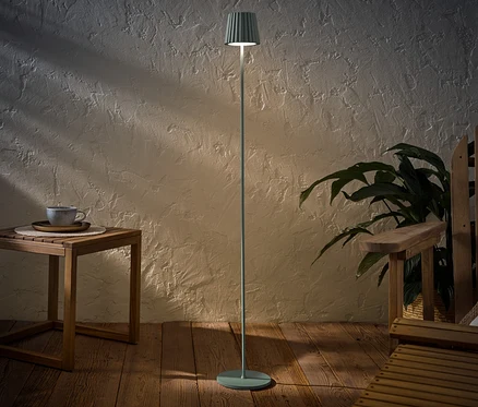 Szałwiowa akumulatorowa lampa stojąca LED do użytku na zewnątrz stoi obok drewnianego stolika z filiżanką i drewnianego krzesła.