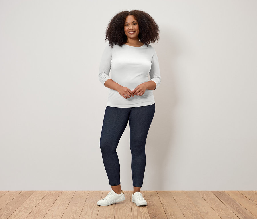 Kobieta z kręconymi włosami stoi na drewnianej podłodze, ubrana w białą koszulkę z rękawami trzy czwarte i ciemnoniebieskie legginsy.