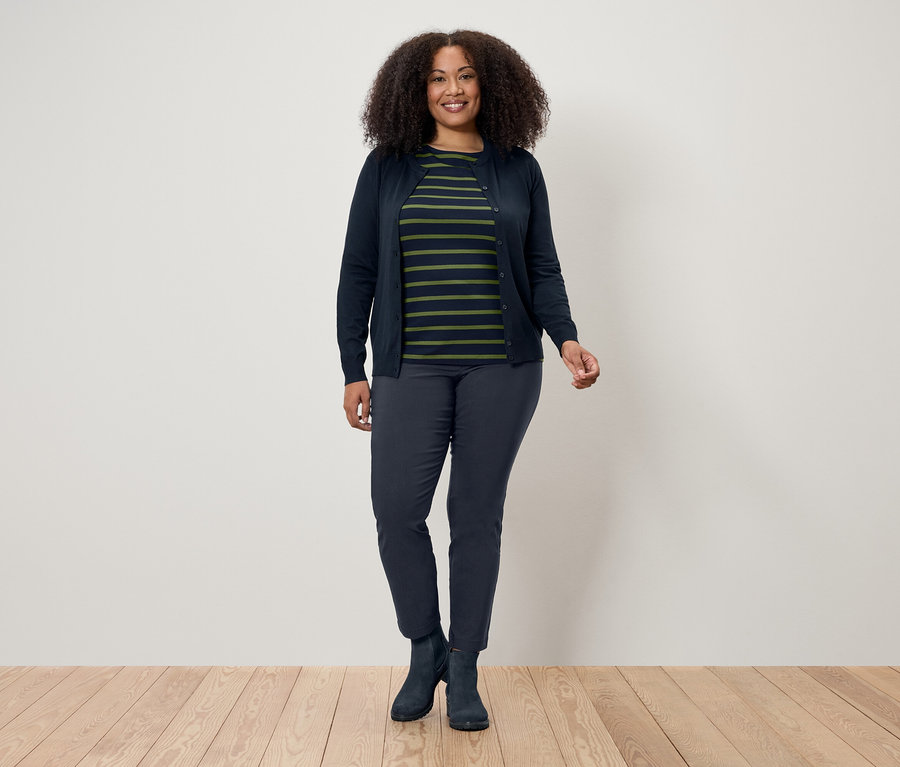 Kobieta z kręconymi włosami stoi na drewnianej podłodze przed białą ścianą. Ma na sobie granatowy kardigan, koszulkę w paski, granatowe spodnie i buty.
