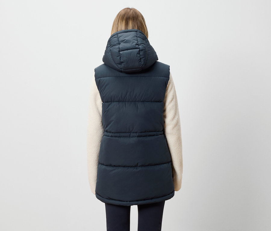Widok z tyłu kobiety z blond włosami, ubranej w granatową pikowaną kamizelkę z kapturem i kremowy sweter.