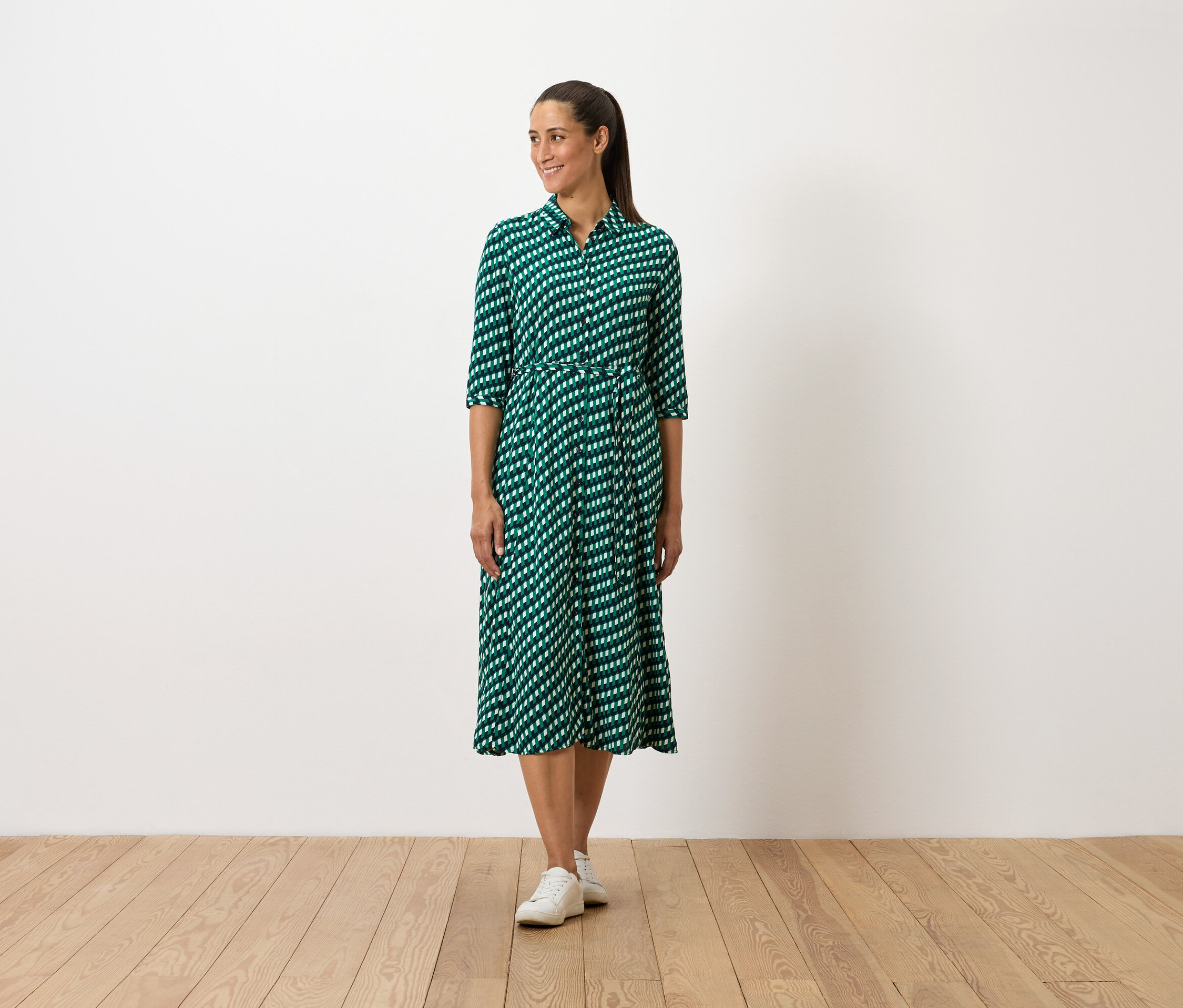 Kobieta stoi w zielonej sukience we wzory i białych tenisówkach.