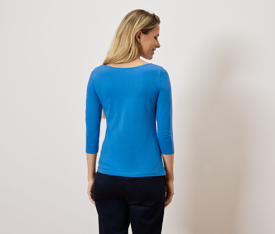 Kobieta patrzy przez ramię, ubrana w niebieską bluzkę z rękawem trzy czwarte i ciemne spodnie.