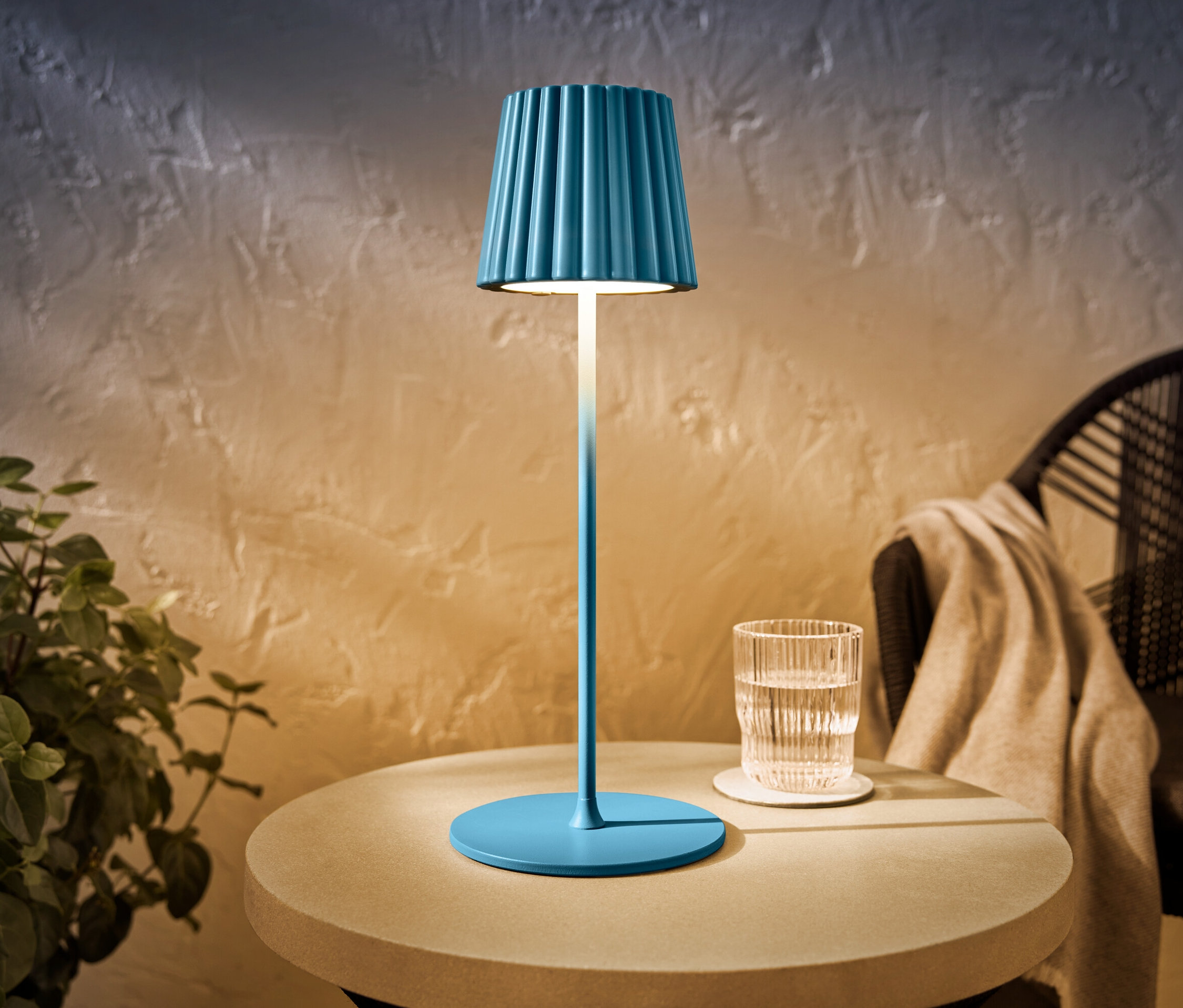 Niebieska akumulatorowa lampa stołowa LED do użytku na zewnątrz stoi na stole obok szklanki wody.