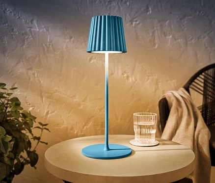 Niebieska akumulatorowa lampa stołowa LED do użytku na zewnątrz stoi na stole obok szklanki wody.