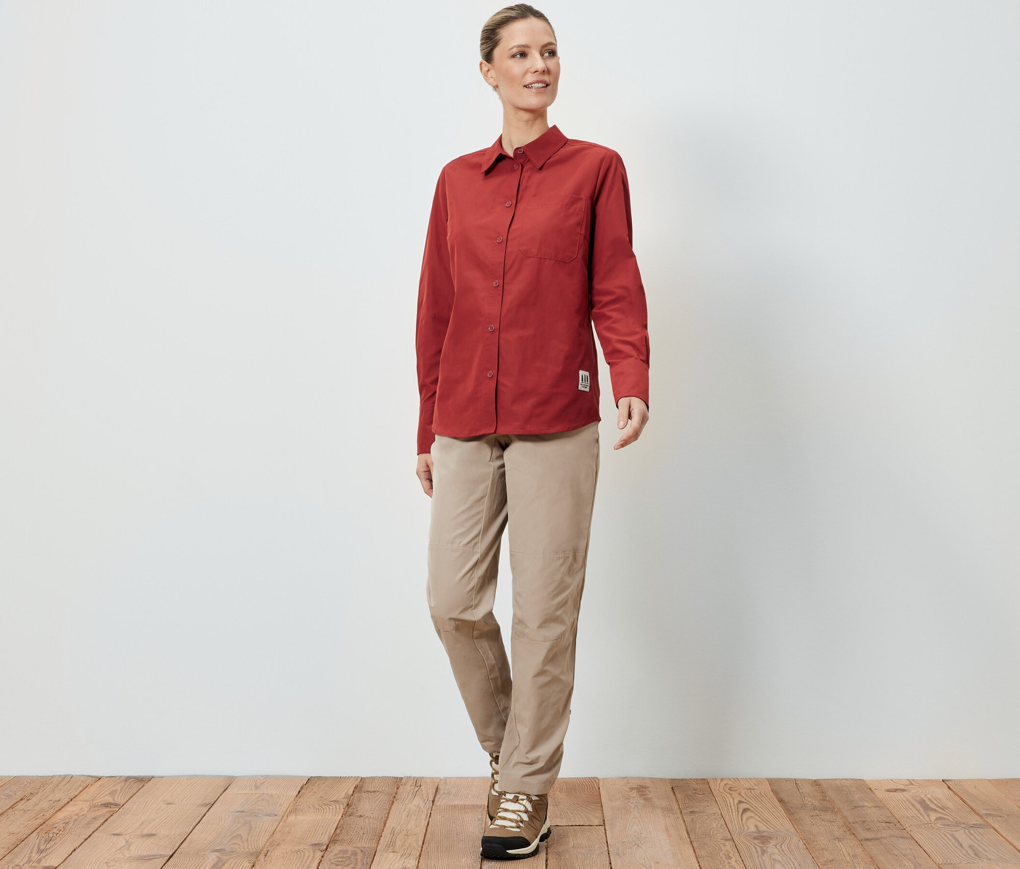 Kobieta w bluzce outdoorowej z ochroną przed komarami, beżowych spodniach i butach turystycznych stoi na drewnianej podłodze przed białą ścianą.