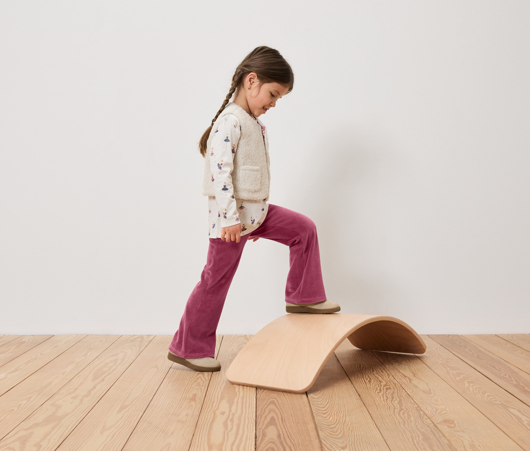 Dziewczynka z warkoczem wchodzi na drewnianą deskę balansującą, ubrana w różowe spodnie sztruksowe, top z nadrukiem i kamizelkę.