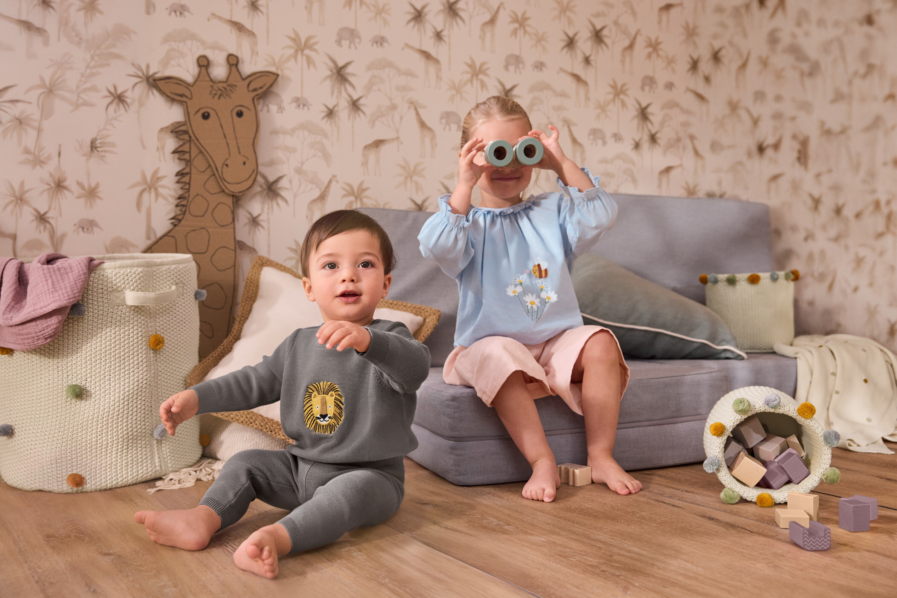 Małe dziecko siedzi obok dużego kosza do przechowywania w pokoju dziecięcym. Dziewczynka siedzi na szarej sofie i trzyma przed twarzą dziergany kocyk dla niemowląt. Ma na sobie dżinsową bluzkę i sztruksowe spodenki typu culottes.