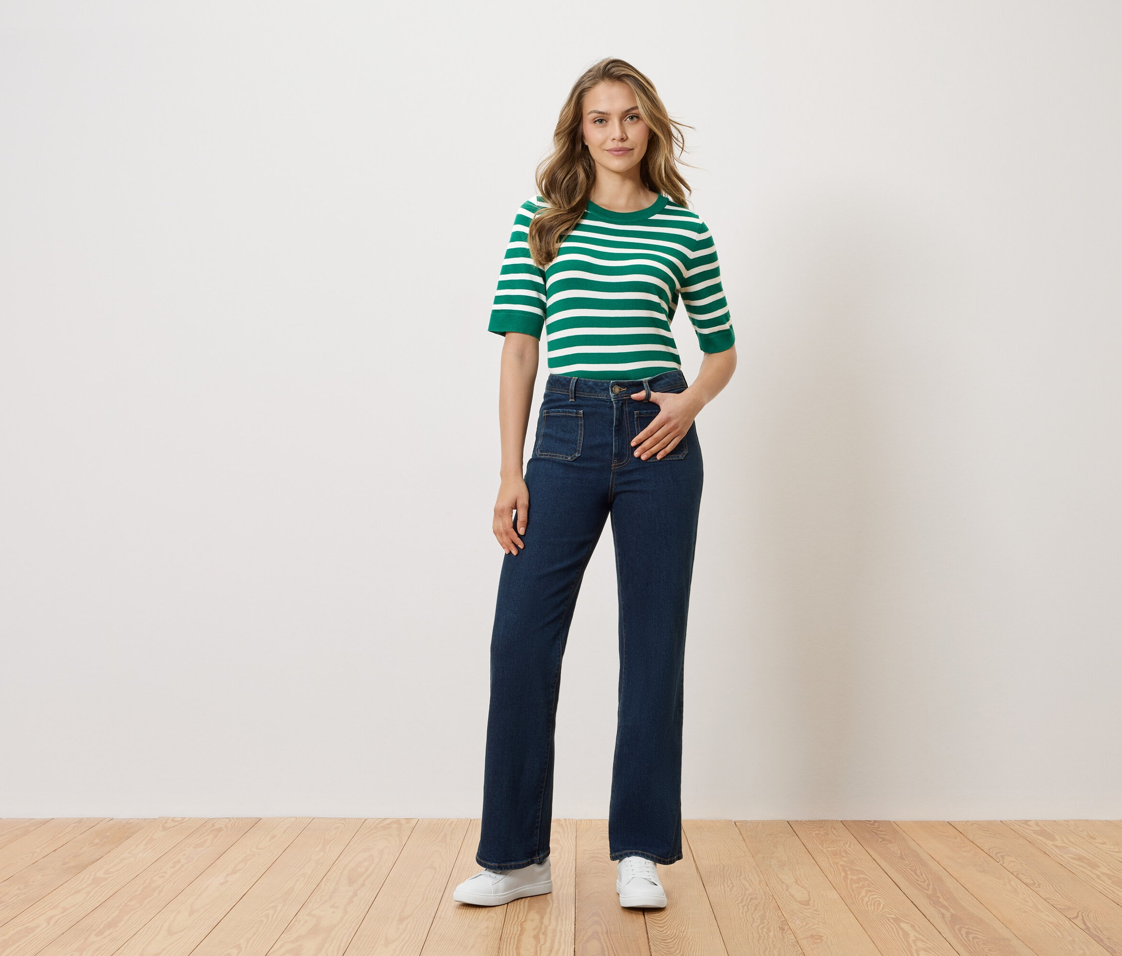 Kobieta stoi z ręką w kieszeni spodni na tle białej ściany. Ma na sobie bluzkę w zielono-białe paski, ciemne dżinsy i białe buty.