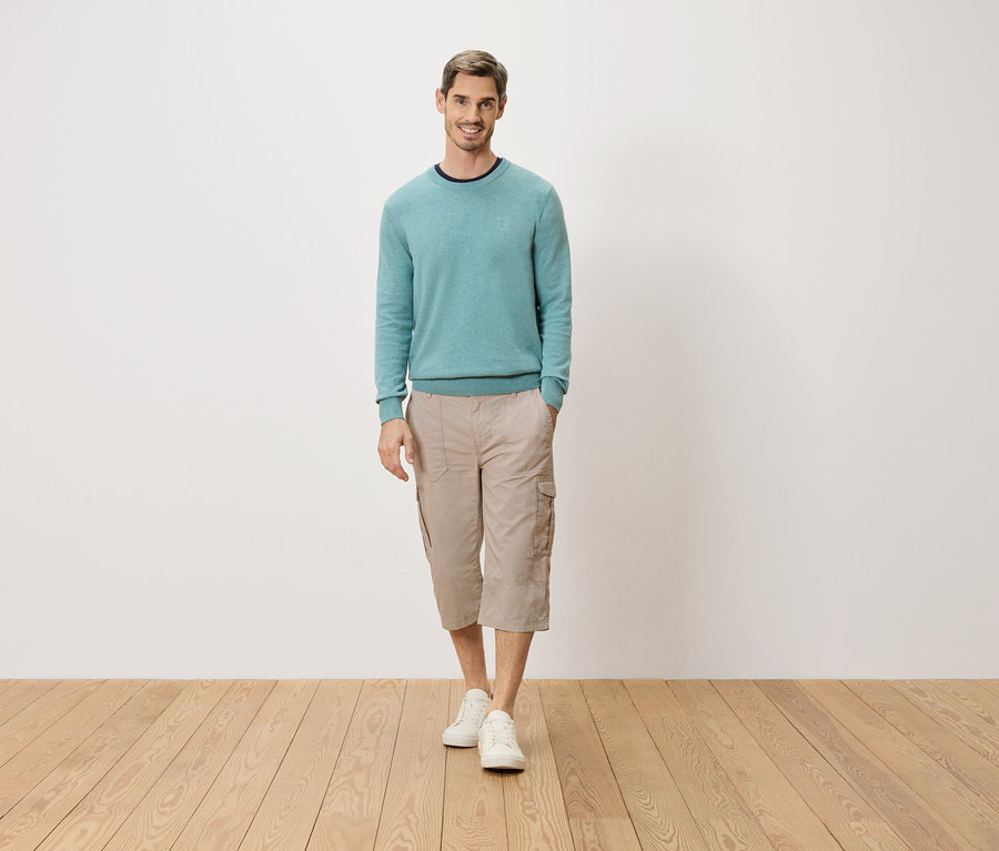 Mężczyzna stoi z ręką w kieszeni na tle białej ściany. Ma na sobie jasnoniebieski sweter, beżowe spodnie cargo 3/4 i białe buty.