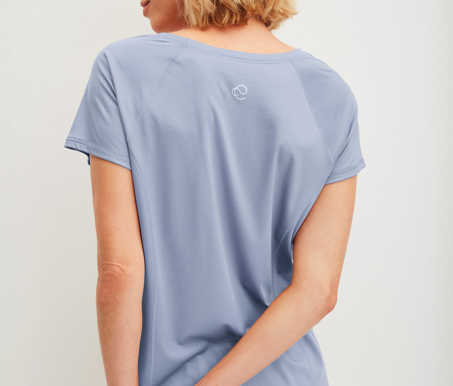 Kobieta z krótkimi blond włosami, widziana od tyłu, ma na sobie jasno błękitny T-shirt z krótkimi rękawami i pasujące krótkie spodenki, ręce splecione za plecami.