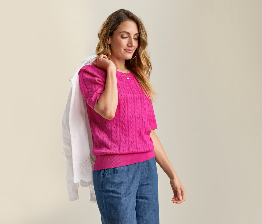 Kobieta ma na ramieniu Jeansjacke mit Lochstickerei i ubrana jest w różowy, cienki sweter z wzorem warkoczowym oraz dżinsowe spodnie.