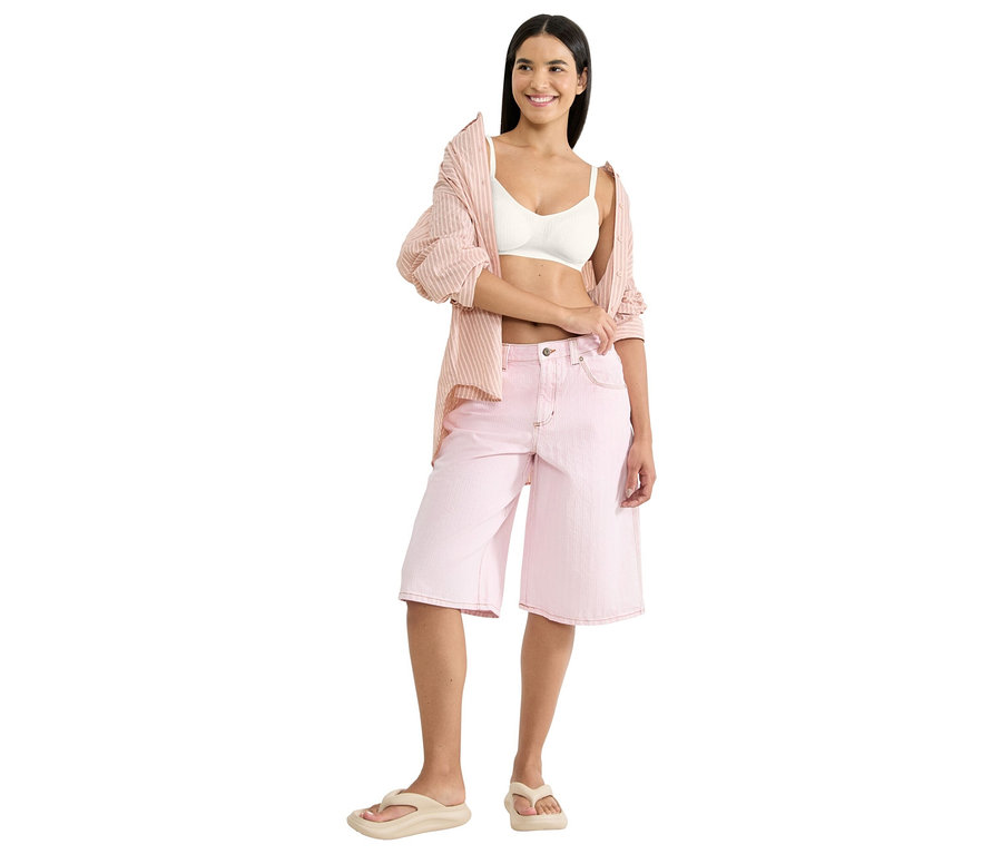 Kobieta ma na sobie biały biustonosz typu bralette, różową koszulę w paski i różowe szorty typu culottes z beżowymi japonkami.