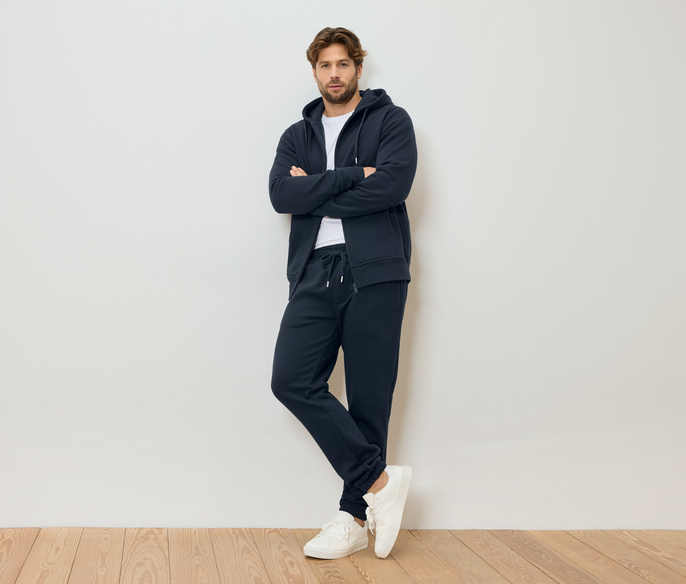 Mężczyzna ze skrzyżowanymi ramionami ma na sobie granatową bluzę z kapturem zapinaną na zamek, granatowe spodnie dresowe i białe tenisówki, stoi na tle białej ściany.
