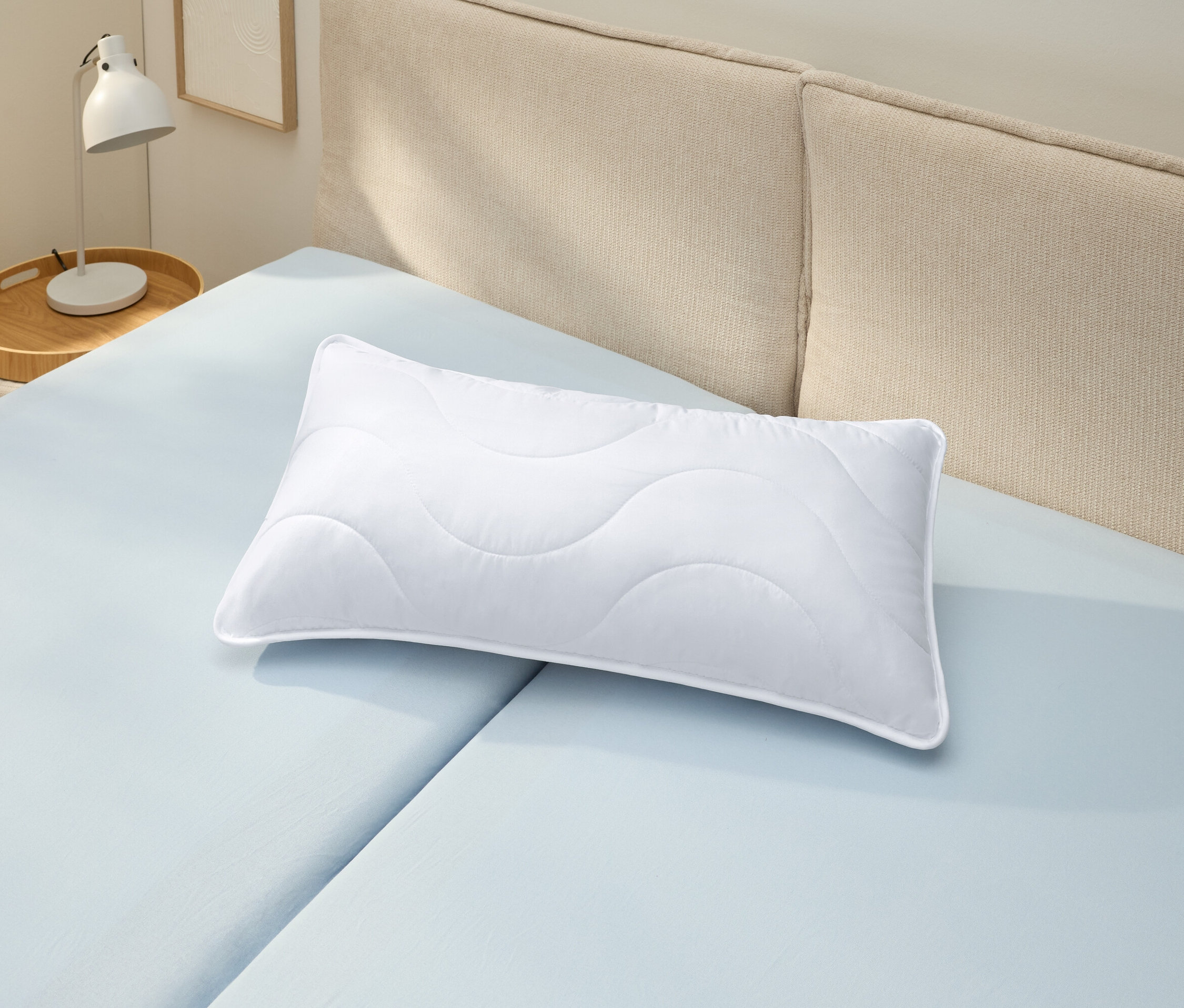 Biała poduszka odporna na pranie w wysokiej temperaturze (ok. 80 x 40) leży na jasnoniebieskim prześcieradle.