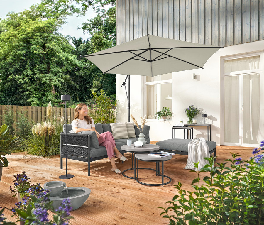 Kobieta siedzi na komfortowej sofie ogrodowej z oddzielnym podnóżkiem. Obok stoi szara solarna lampa podłogowa. Na pierwszym planie solarna fontanna ogrodowa.