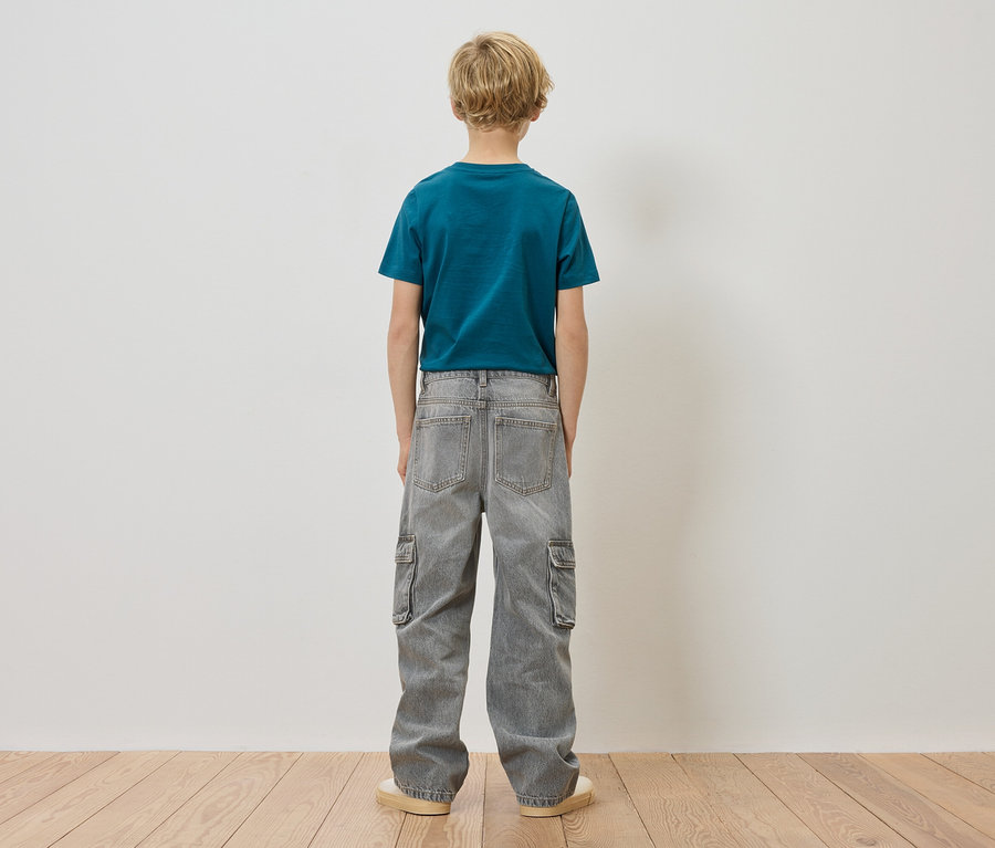 Chłopiec o blond włosach stoi tyłem do kamery, ubrany w niebiesko-zielony T-shirt i szare dżinsy cargo.