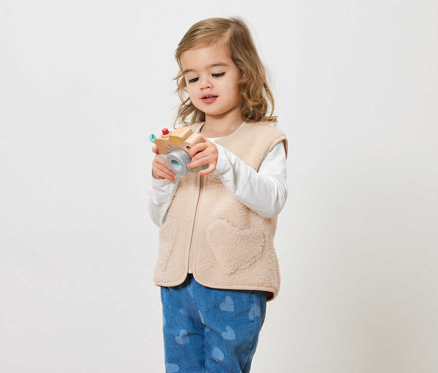Mała dziewczynka z kręconymi włosami trzyma zabawkę-aparat na jasnym tle.