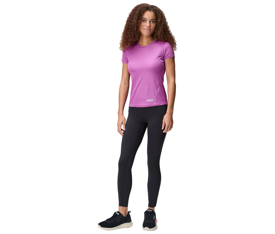 Kobieta stoi, ma na sobie fioletową koszulkę Fila, czarne legginsy i czarne buty sportowe.