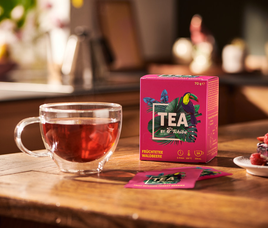 Na drewnianym stole stoi szklanka z herbatą owocową, opakowanie herbaty TEA by Tchibo o smaku leśnych jagód i talerz z deserem.