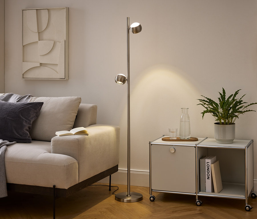 Nowoczesny salon z beżową sofą, lampą podłogową ze szczotkowanego metalu i modułową szafką. Na sofie leży szary koc i poduszka, a na podłokietniku otwarta książka. Lampa oświetla obszar obok szafki z rośliną, karafką wody i książkami.
