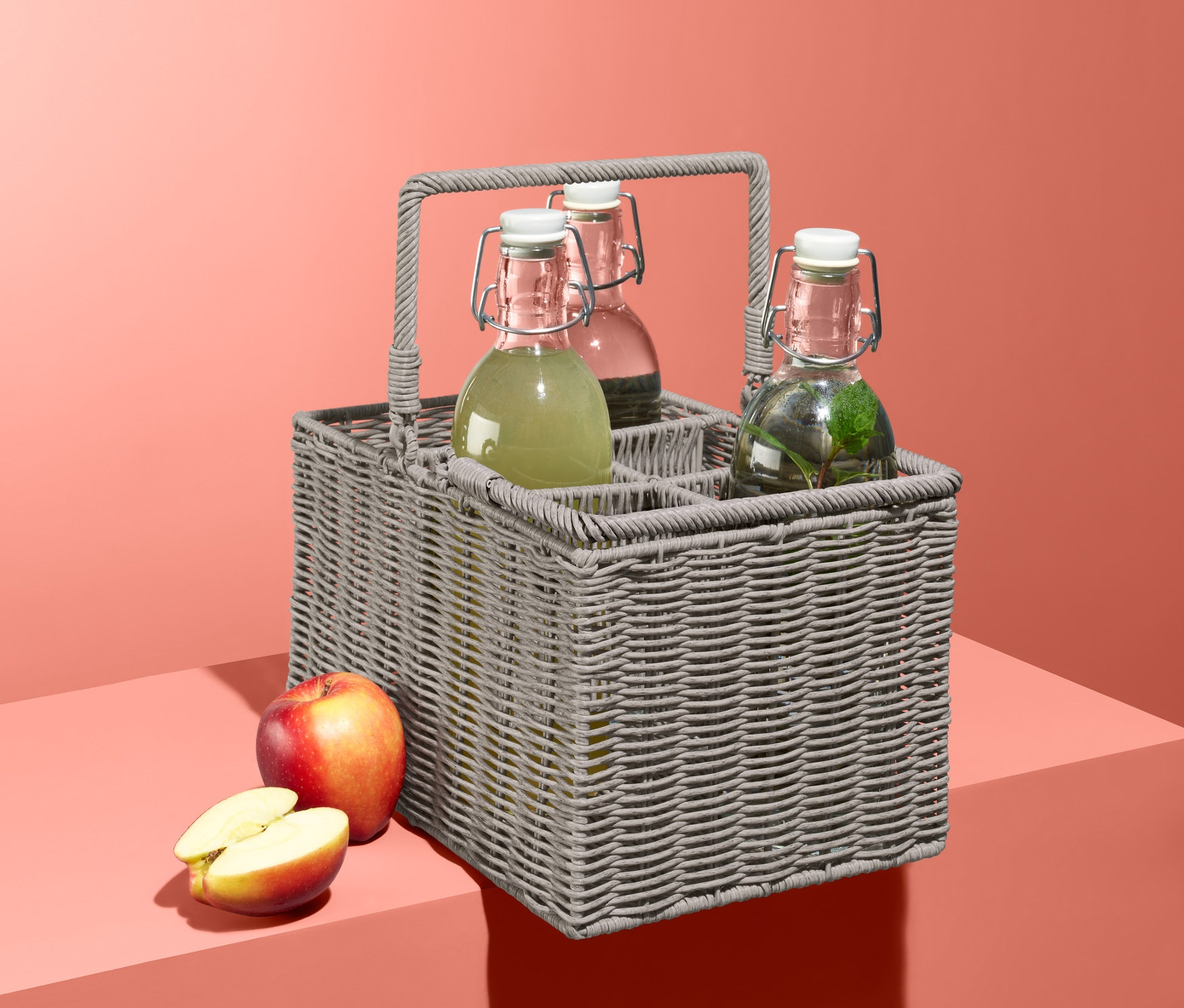 Szary kosz na butelki z trzema szklanymi butelkami i jabłkiem na koralowym tle.