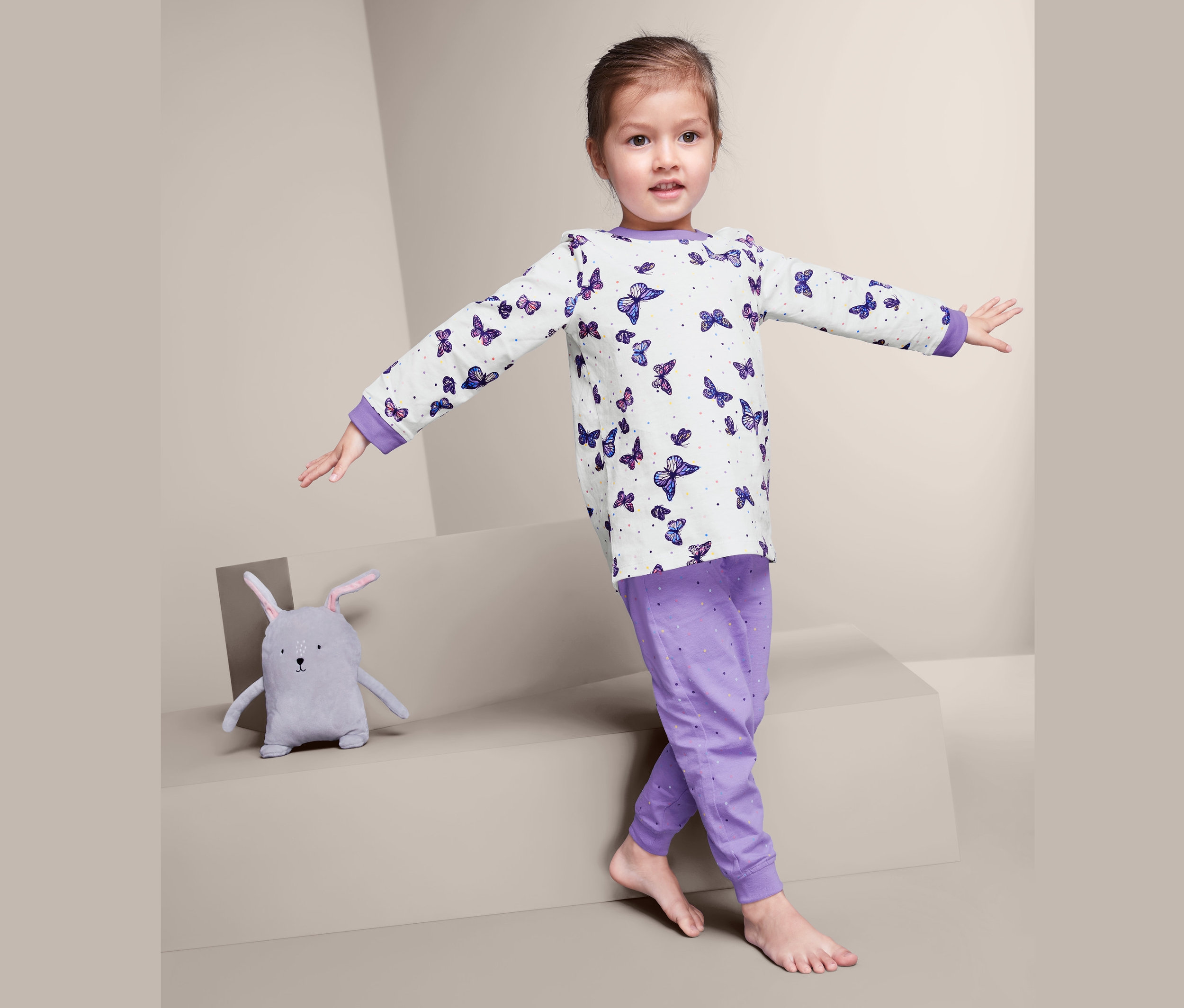 Dziewczynka w piżamie w motyle stoi z rozłożonymi ramionami obok pluszowego króliczka.