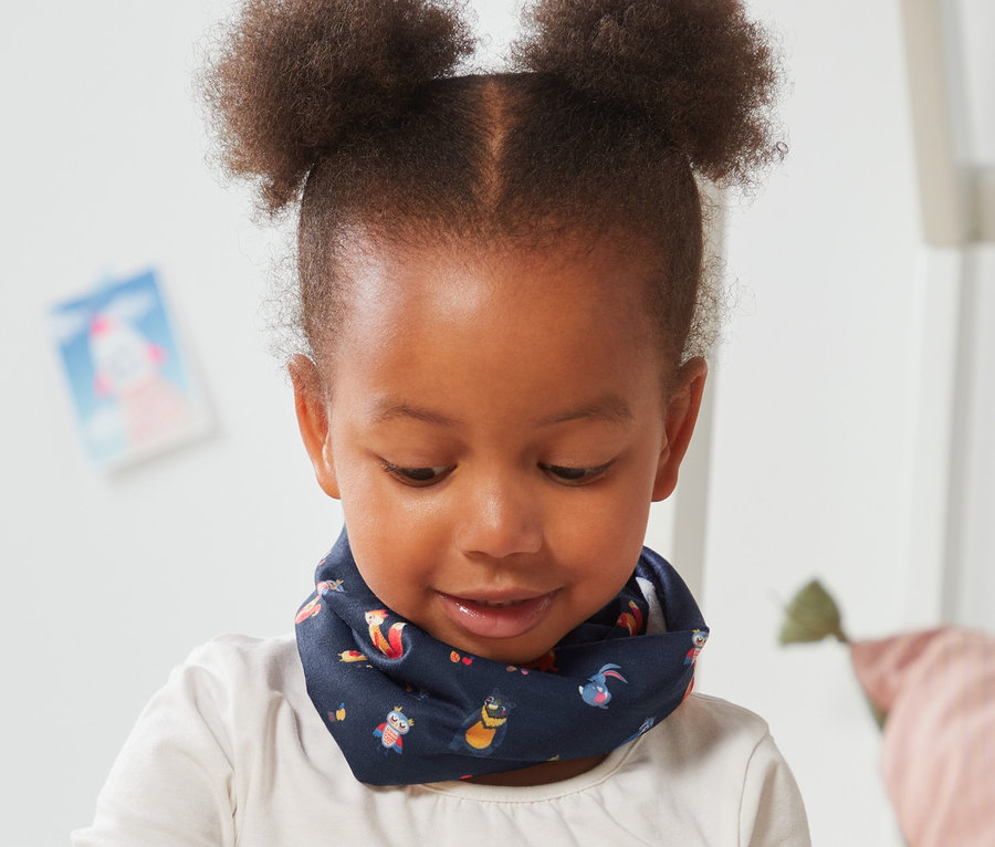 Dziewczynka siedzi na podłodze i bawi się drewnianymi klockami. Ma na sobie chustkę wielofunkcyjną z nadrukiem na całej powierzchni.