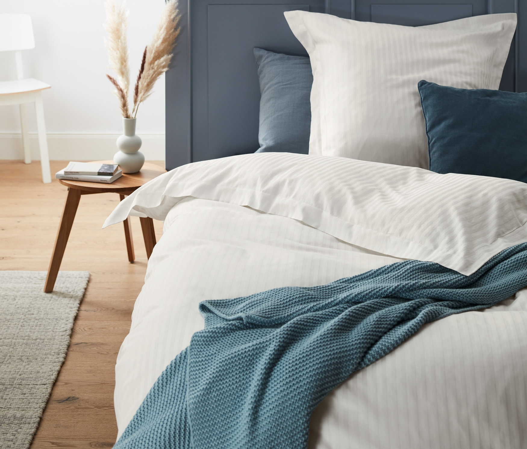 Kremowa satynowa pościel premium na łóżku z niebieskim kocem i poduszkami.