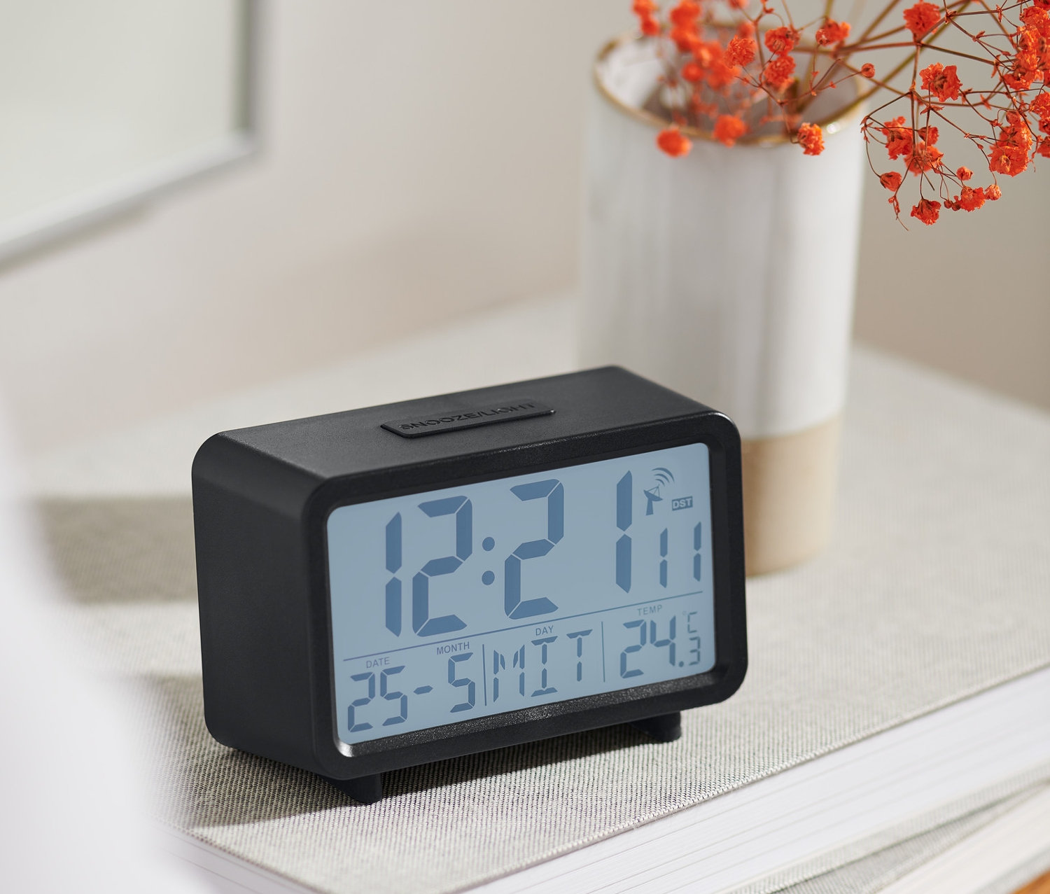 Czarny budzik cyfrowy sterowany sygnałem radiowym z wyświetlaczem LCD stoi na książce obok wazonu z pomarańczowymi kwiatami.