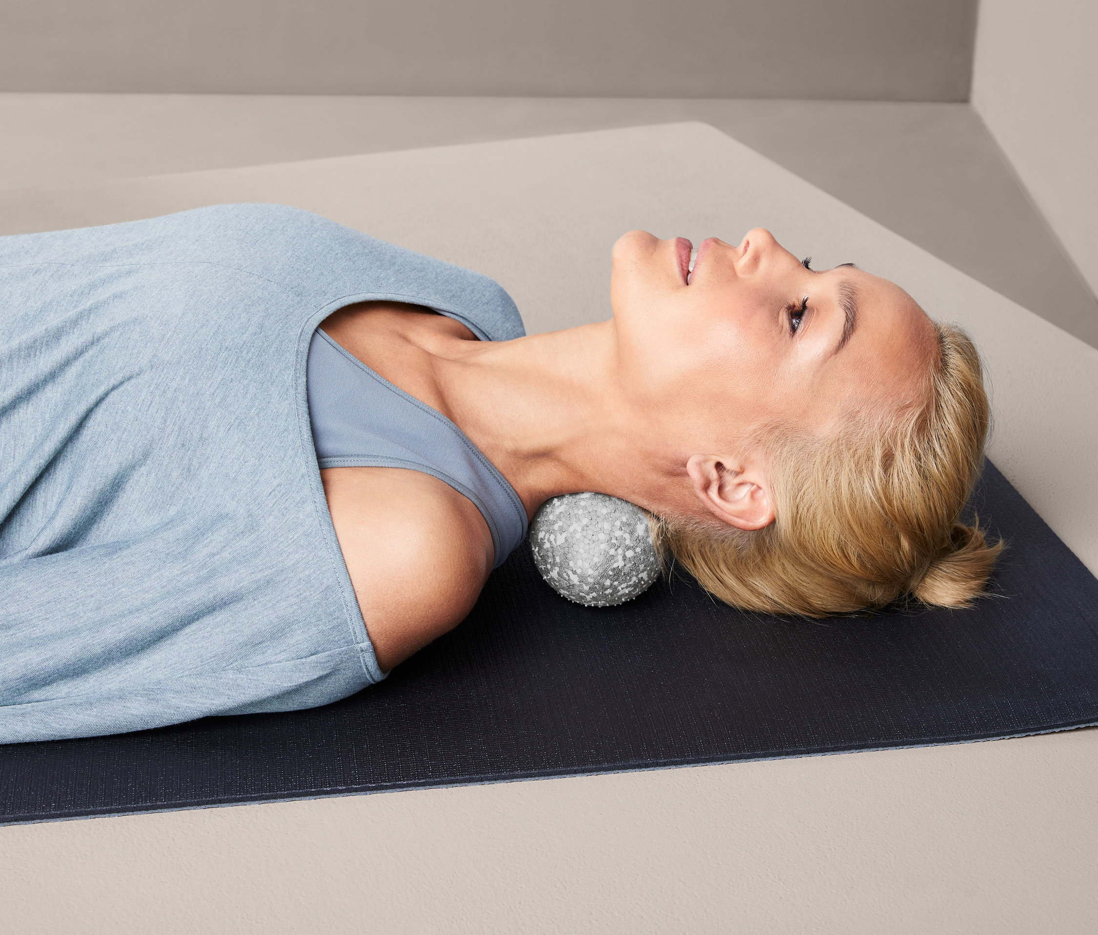 Kobieta leży na macie z Podwójną piłką do masażu powięzi pod szyją.
