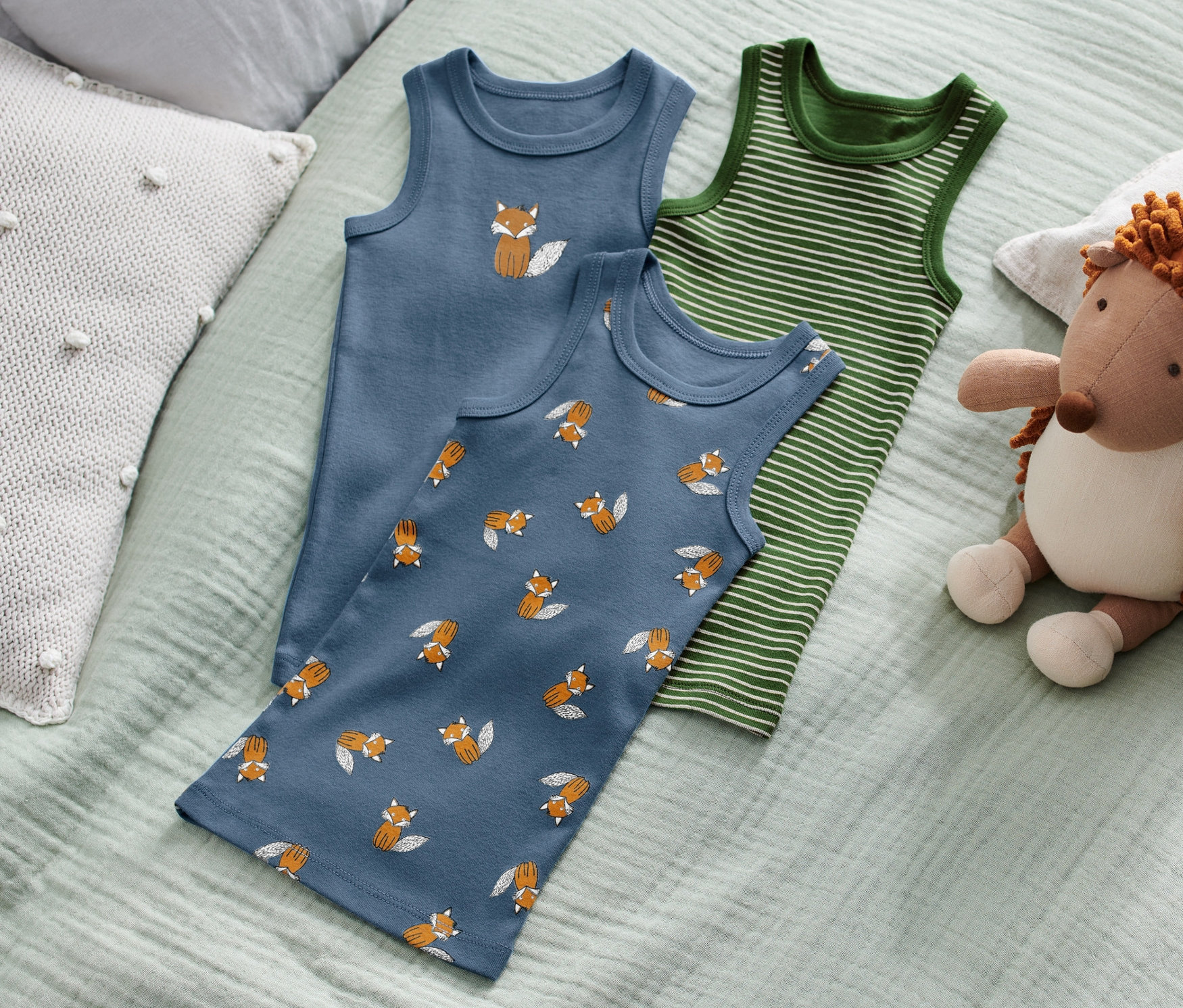 Na łóżku leżą trzy podkoszulki dziecięce z motywem lisa oraz pluszowy jeż.