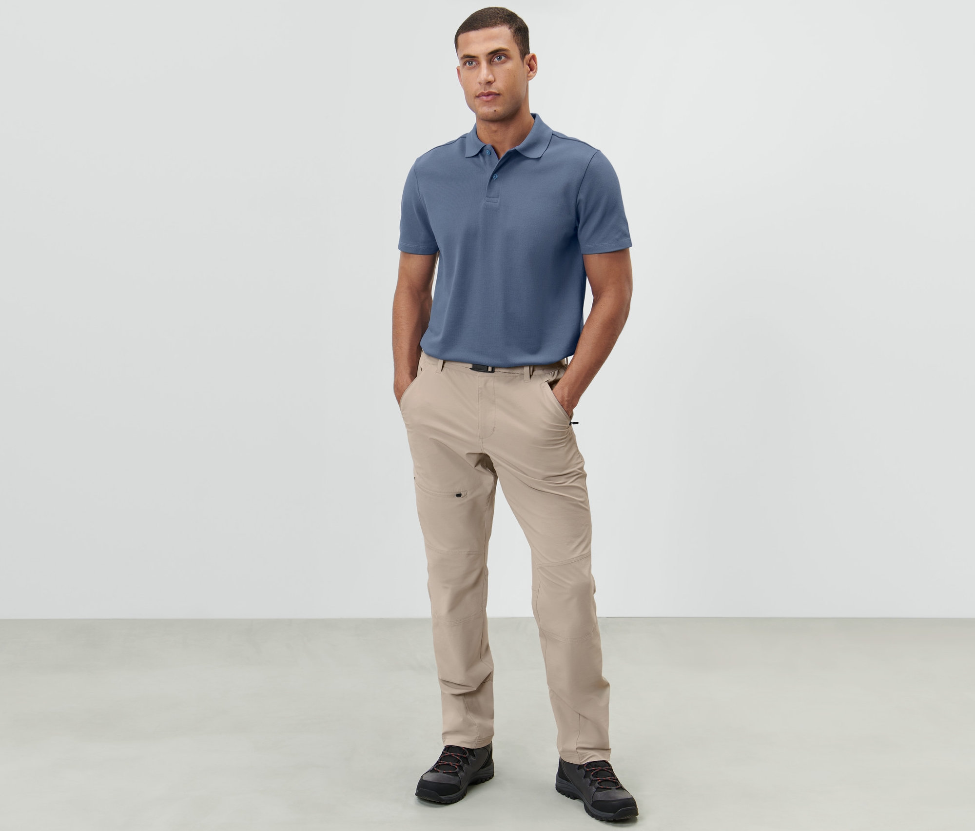 Mężczyzna stoi z rękami w kieszeniach jasnobeżowych spodni outdoorowych.