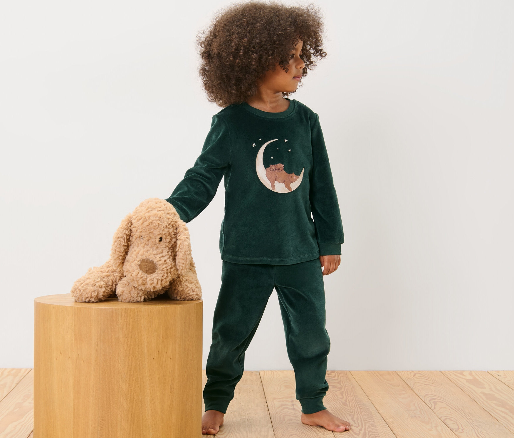 Dziecko w piżamie z weluru stoi obok pluszowego psa na drewnianym stołku.