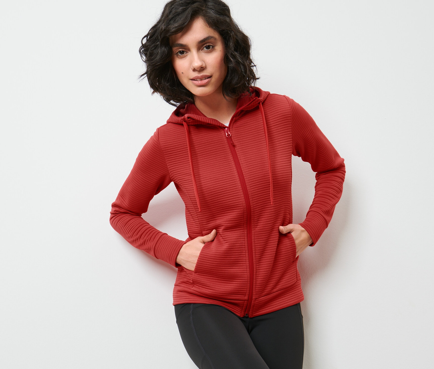 Kobieta pozuje w czerwonej bluzie z kapturem i czarnych legginsach.