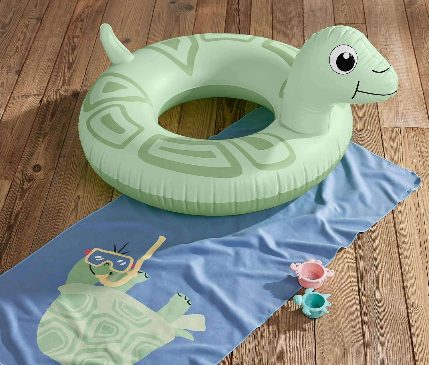 Zielone kółko do pływania „Żółw” leży na niebieskim ręczniku z nadrukiem żółwia i dwiema zabawkami na drewnianej podłodze.
