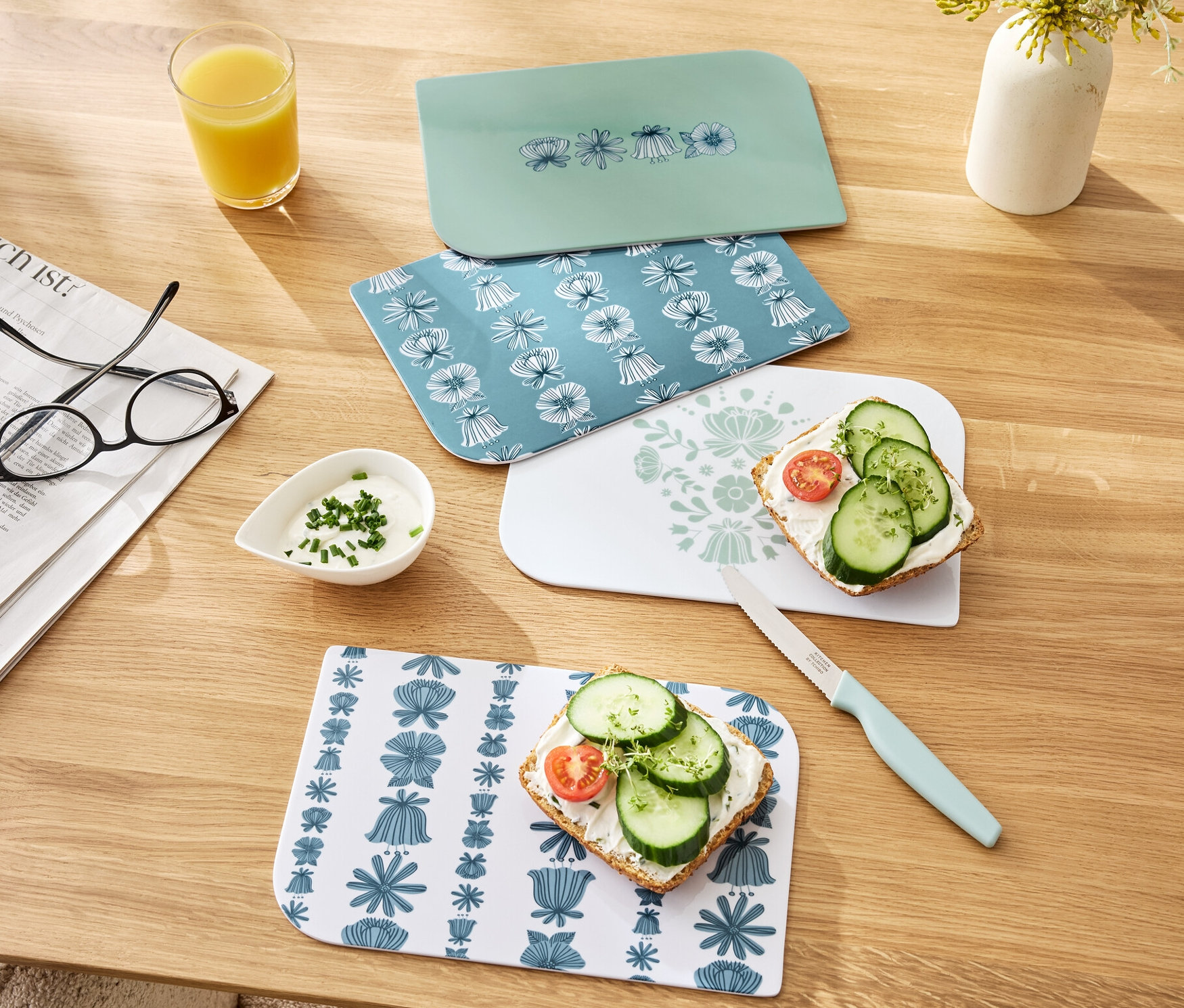 Cztery deski śniadaniowe leżą na drewnianym stole, dwie z kanapkami. Widoczne są również szklanka soku pomarańczowego, miska z dipem i nóż.