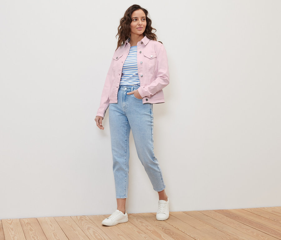 Kobieta pozuje w różowej kurtce dżinsowej i jasno niebieskich dżinsach.