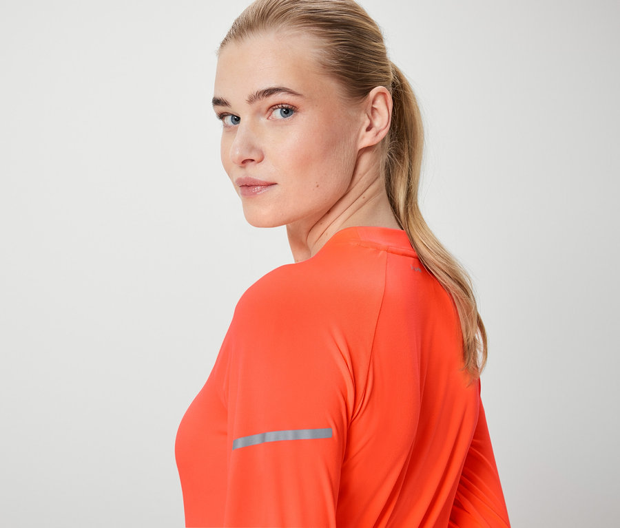 Blondwłosa kobieta spogląda przez ramię, ubrana w pomarańczową bluzkę z długim rękawem.
