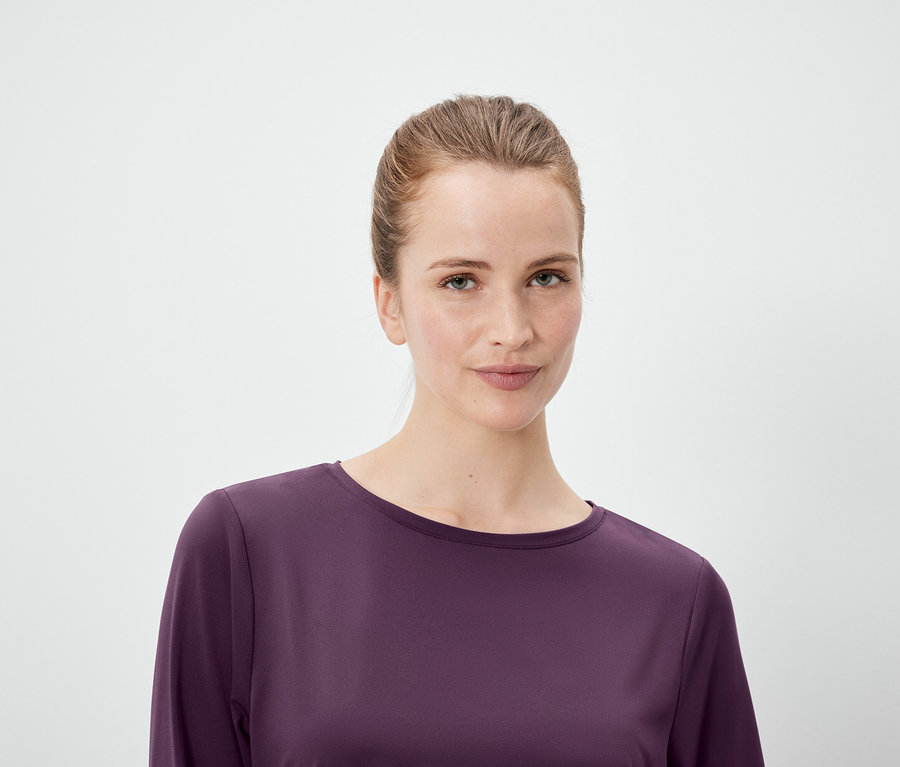 Portret kobiety z włosami związanymi w koński ogon, ubranej w fioletową bluzkę z długim rękawem na białym tle.