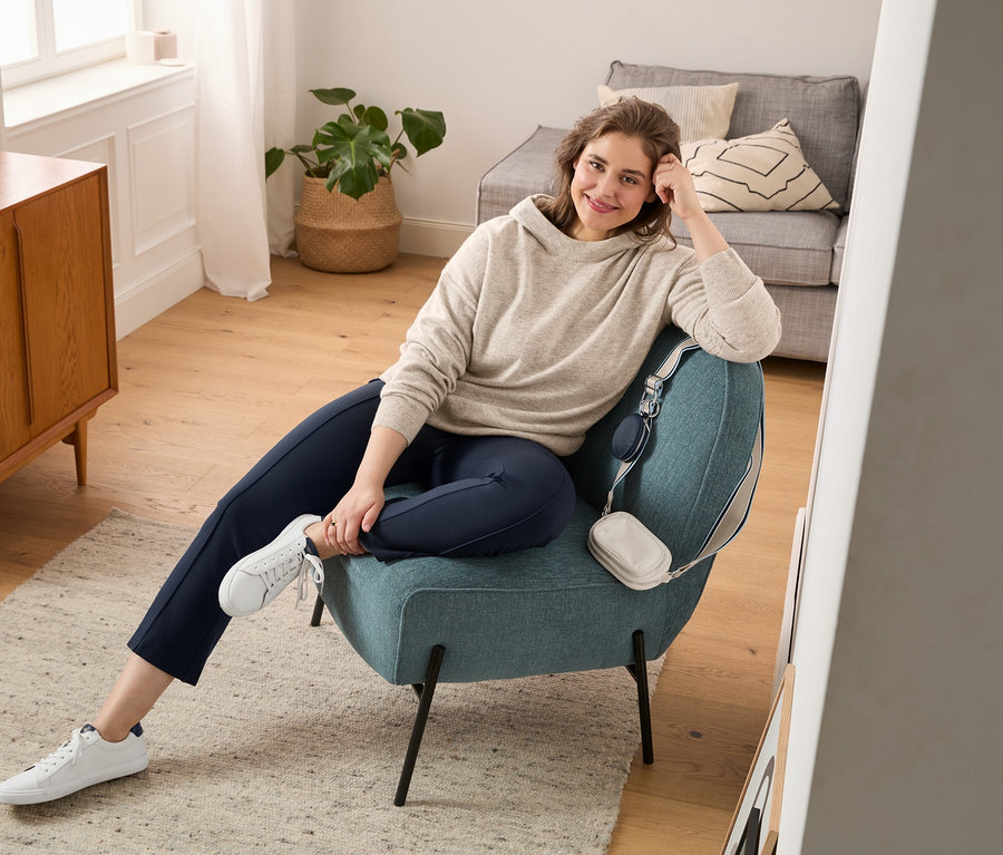 Kobieta siedzi w fotelu, ma na sobie sweter z delikatnej dzianiny z kaszmirem, rozszerzane legginsy i buty typu sneaker ze skóry garbowanej bezchromowo. Na krześle wisi torba na ramię do noszenia na skos.