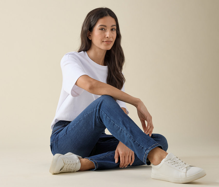 Kobieta siedzi na podłodze w dżinsach slim- fit »Emma« w kolorze mid blue denim, białym t-shircie i sneakersach.