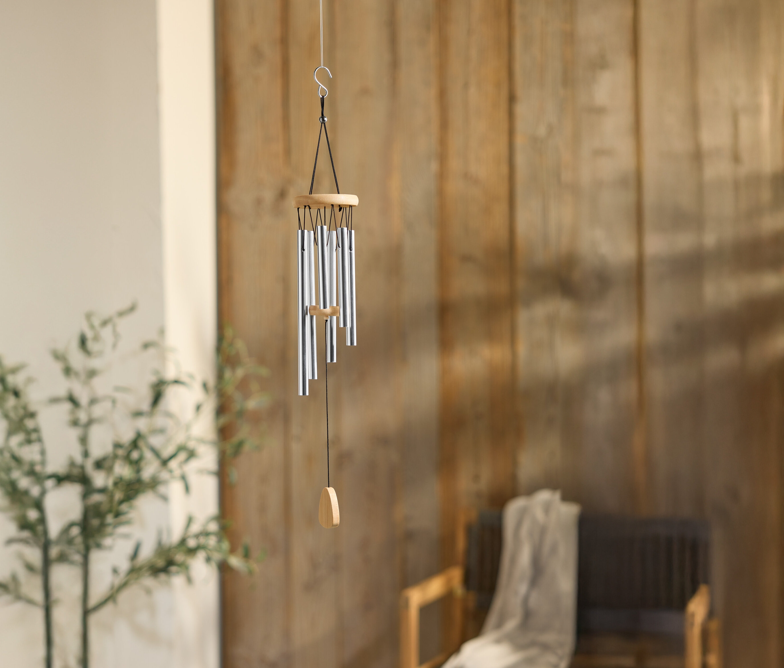 Dzwonek wietrzny z srebrnymi rurkami wisi na drewnianej ścianie, obok rośliny i krzesła.