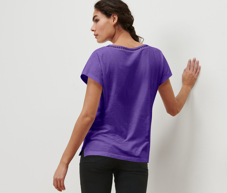 Kobieta z zaplecionymi włosami stoi tyłem, opierając się o ścianę, ma na sobie liliową bluzkę z mieszanki modalu.
